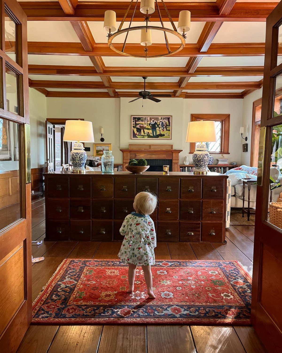 Rustic living room with wood beams, red rug, and a child standing nearby.