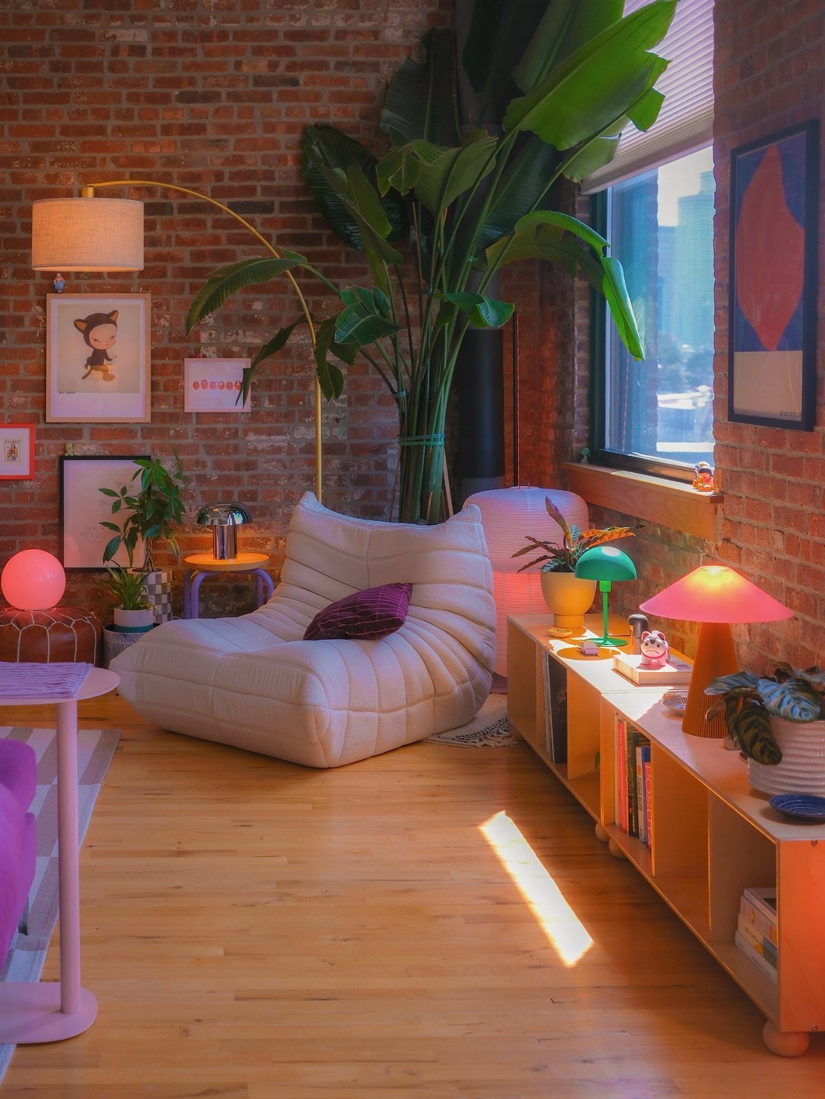 Living room with white lounge chair and colorful lamps against a brick wall.