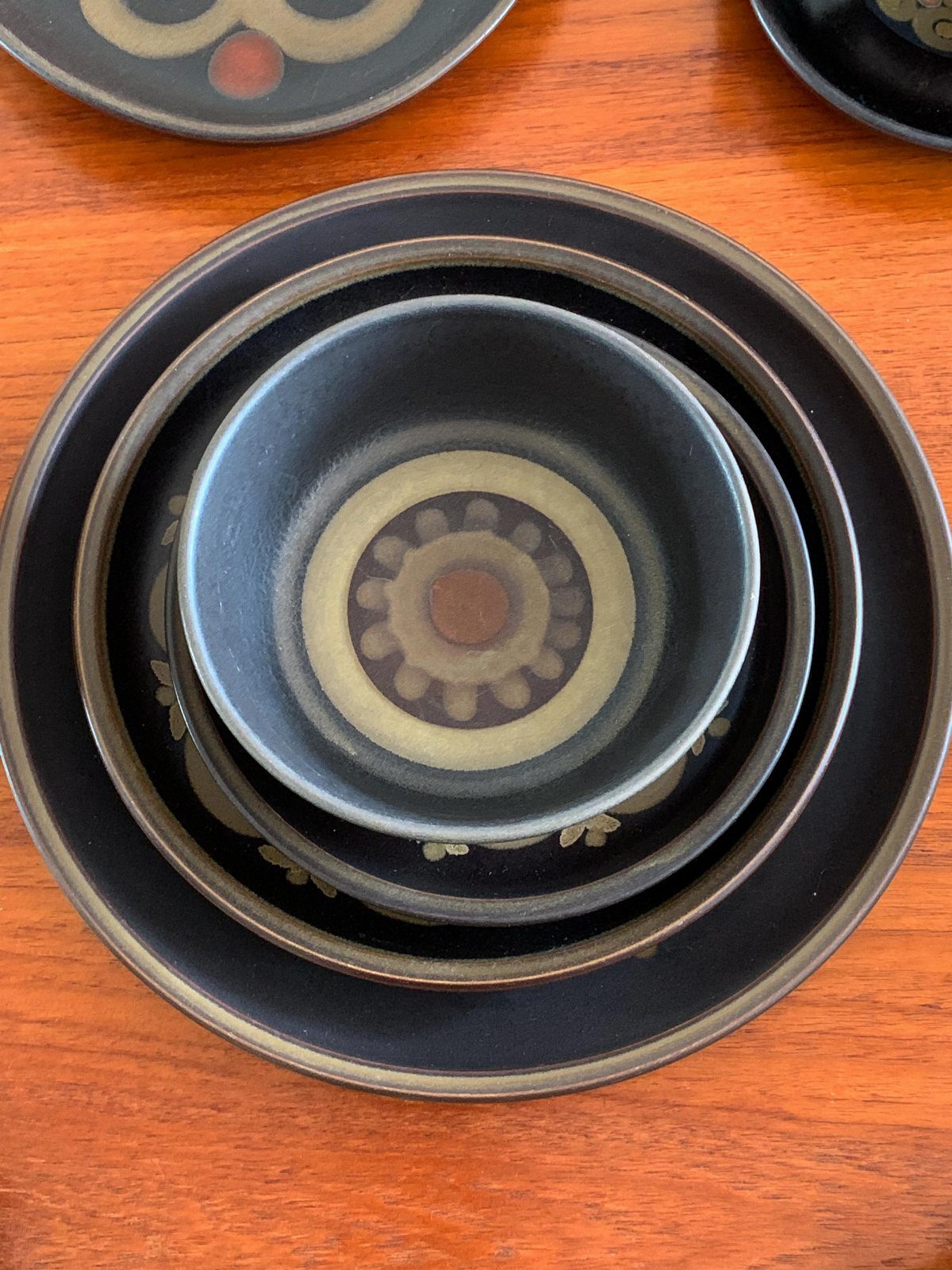 Denby Dinnerware Sets in 5 Classic Styles 6 Stacked dark brown ceramic plates and bowl with circular patterns on a wooden table.