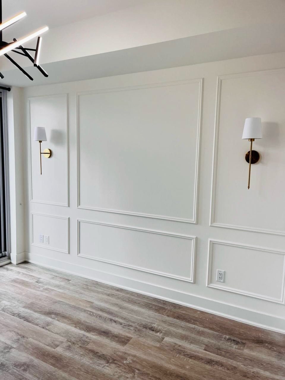 White wall with decorative architectural moldings, gold sconces, and light wood flooring.