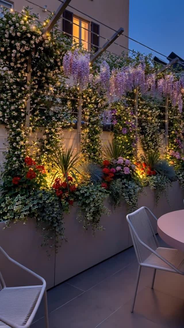 Lush balcony garden with purple wisteria, red flowers, and warm fairy lights at dusk.