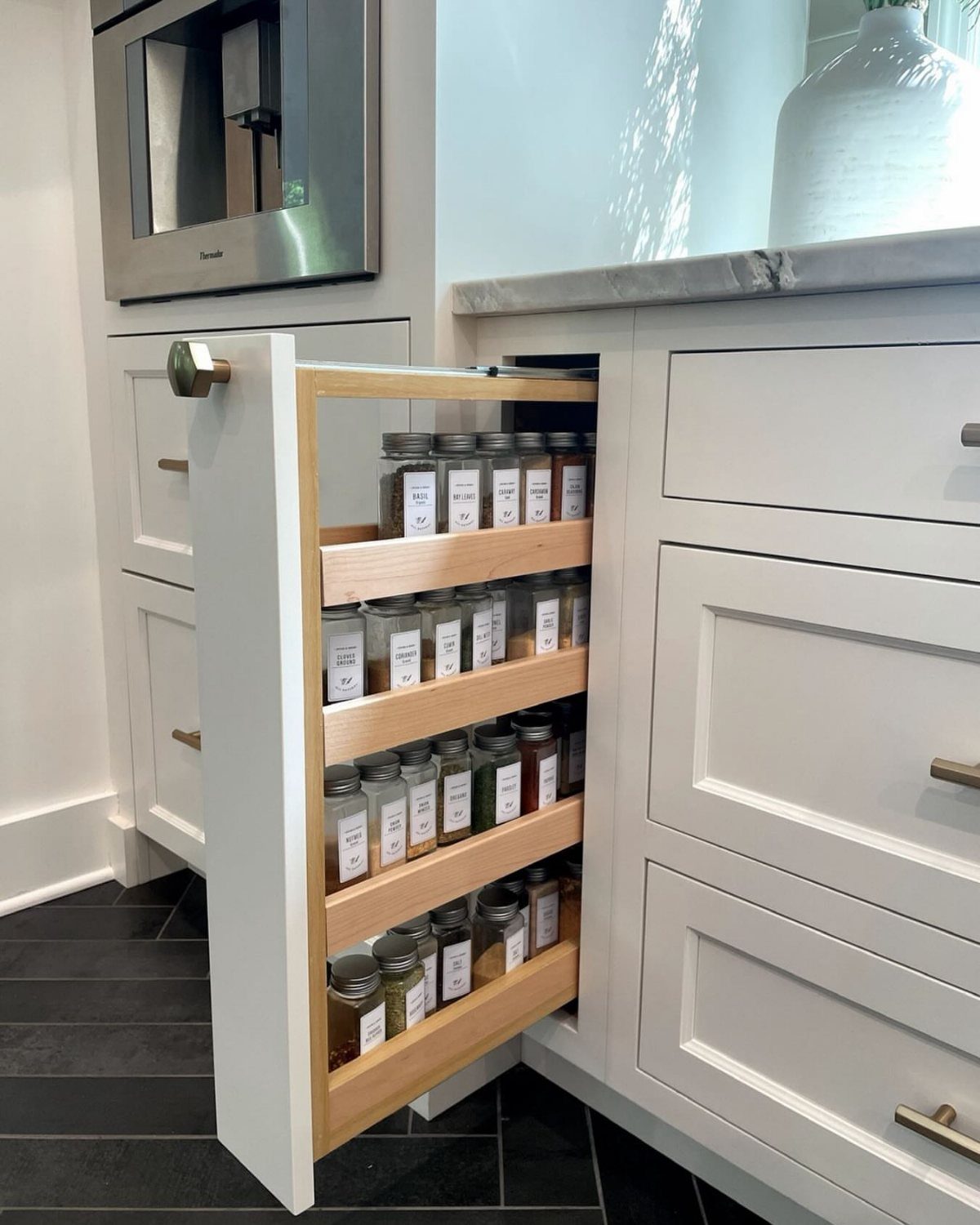 Pull-out wooden spice rack with glass jars extending from white kitchen cabinetry.