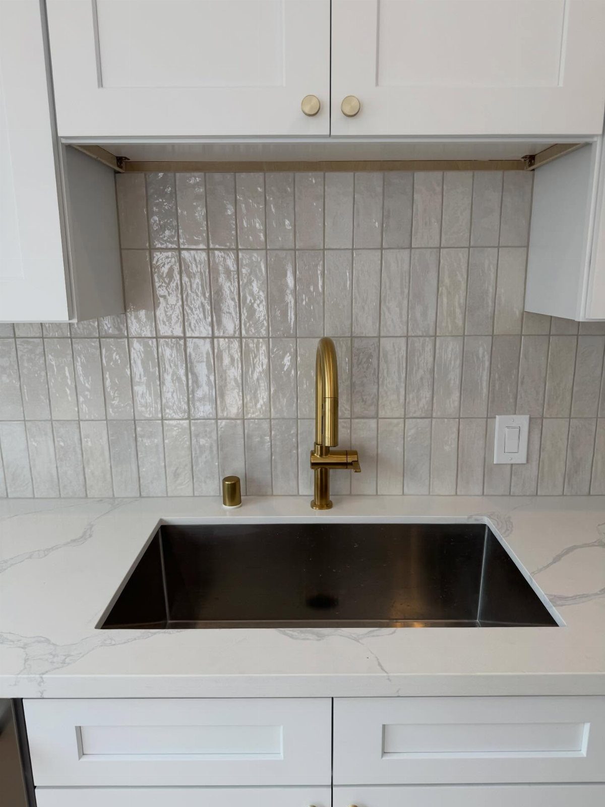 Light gray vertical tile backsplash behind a gold faucet and stainless steel sink.