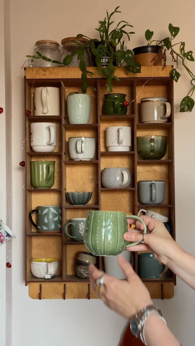 Wooden wall grid organizer holding colorful ceramic mugs with a hand reaching for one.