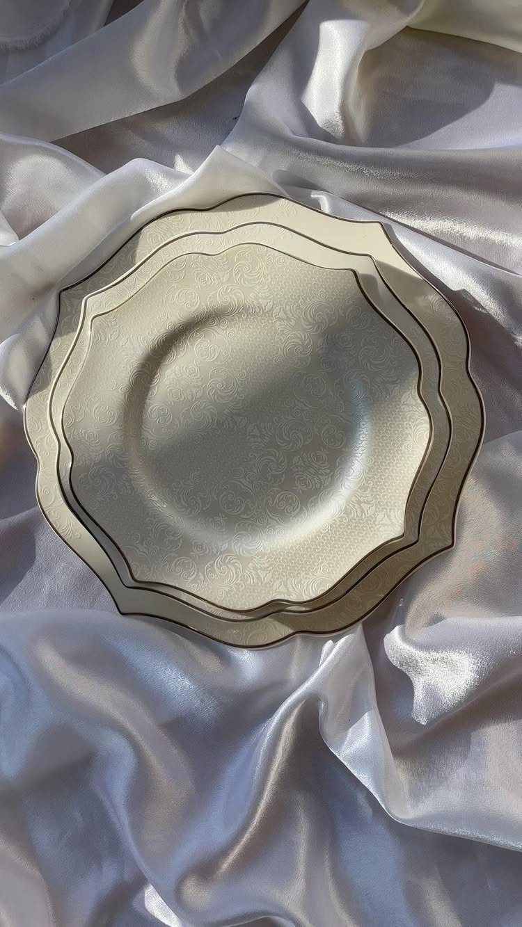 Fancy Dinnerware Set Ideas for 2026 Tables (28 Styles) 20 Cream-colored scalloped dinner plates stacked on draped white satin fabric.