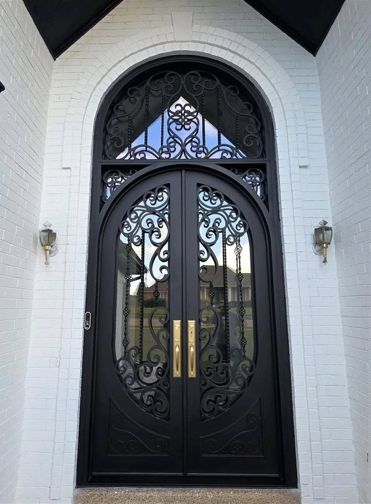 Ornate black double front door with wrought iron scrollwork in white brick archway.