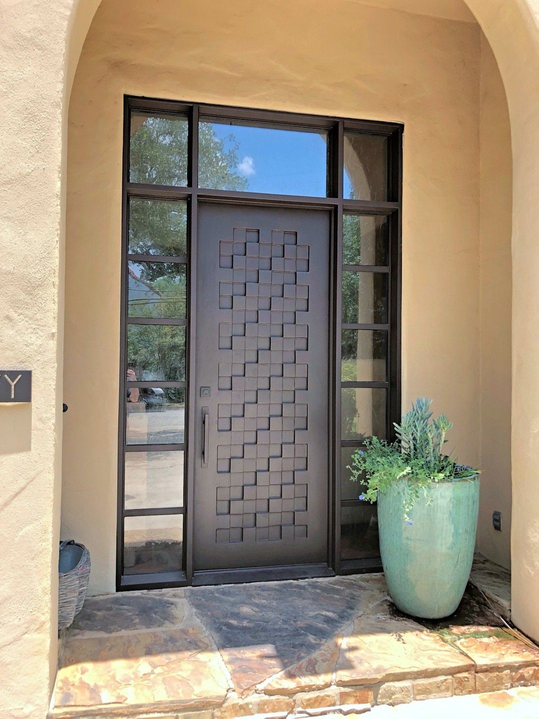 Modern dark metal front door with geometric patterns in a tan stucco entryway.
