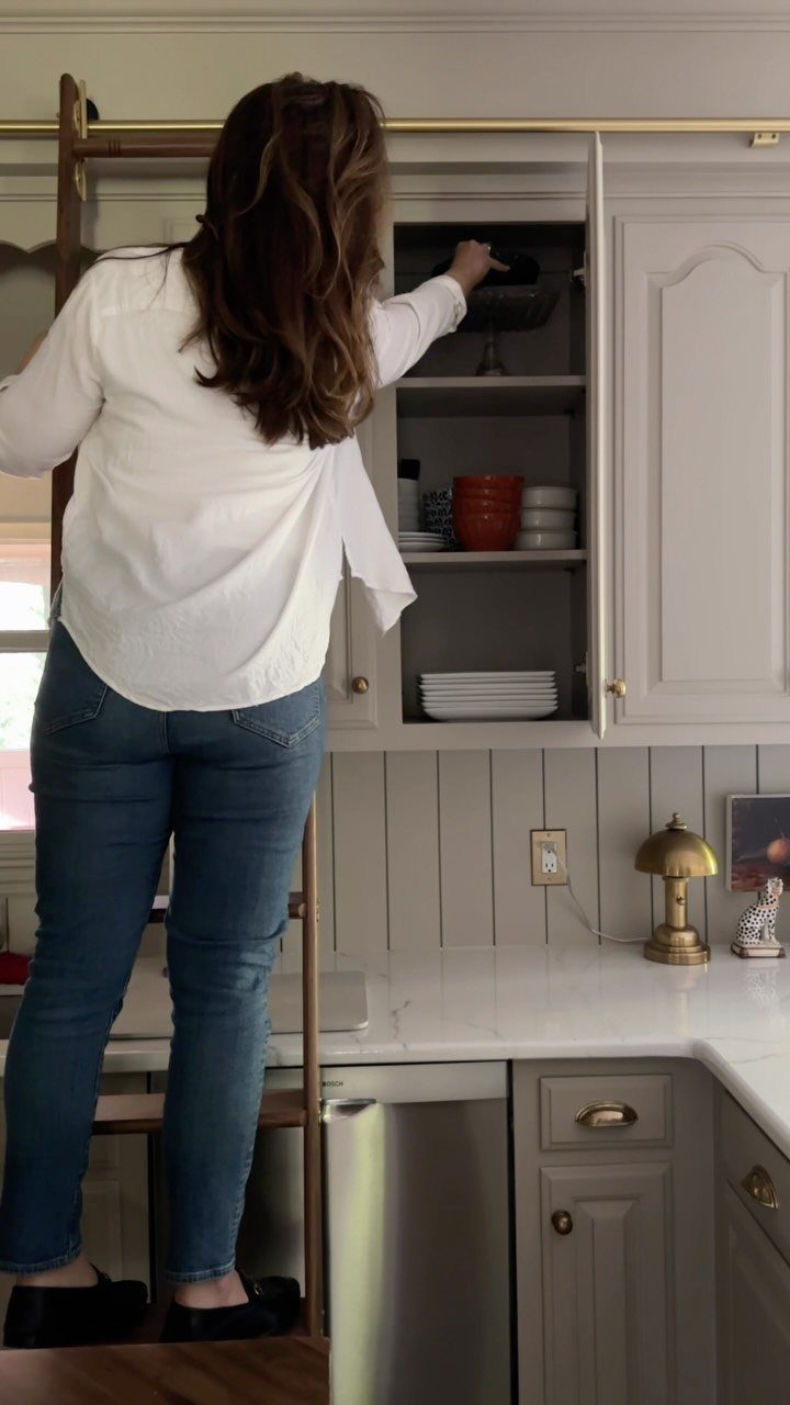Woman on a wooden ladder reaching into an open grey kitchen cabinet