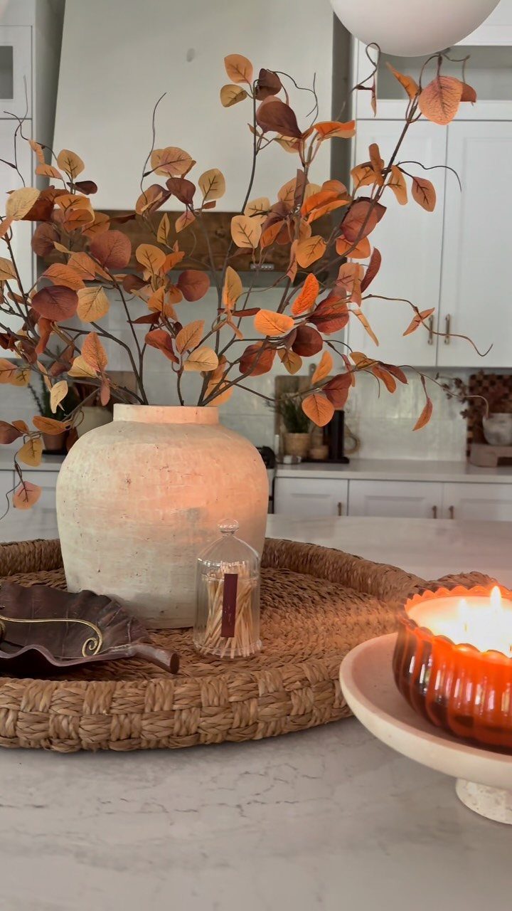 14 Kitchen Tabletop Decor Ideas to Style Your Counters 13 Lit orange ribbed candle on a white pedestal next to a vase of autumn leaves.