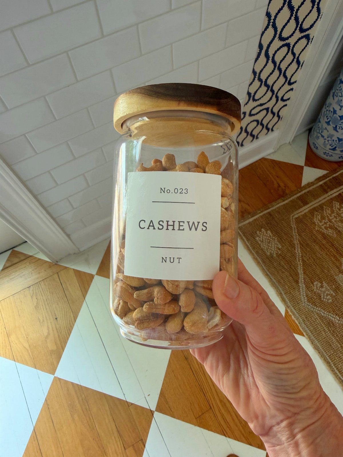 14 Kitchen Tabletop Decor Ideas to Style Your Counters 10 Glass jar with wooden lid filled with cashews held against white tiled wall.