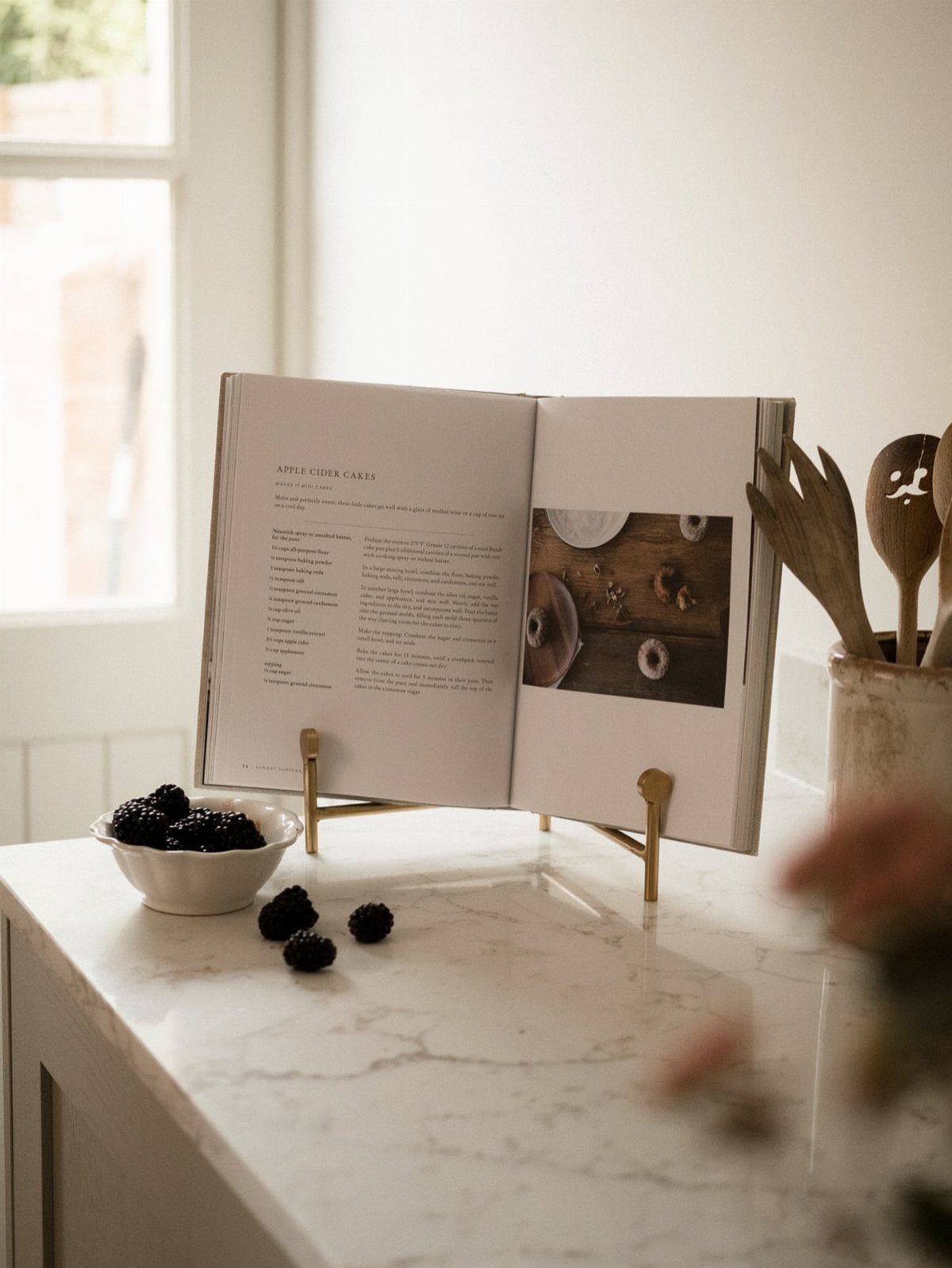 14 Kitchen Tabletop Decor Ideas to Style Your Counters 7 Open cookbook on a gold stand next to blackberries on a marble countertop