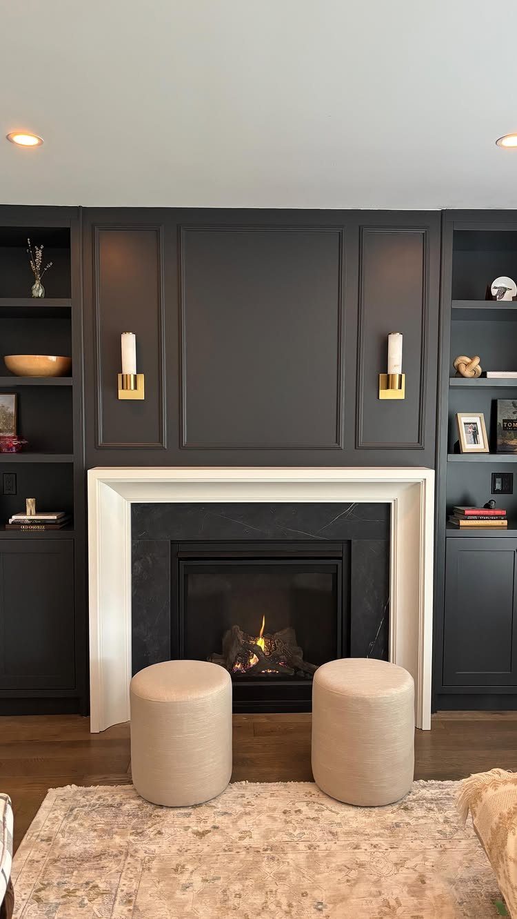 Dark grey built-in cabinets flanking a white mantel fireplace with two beige ottomans.