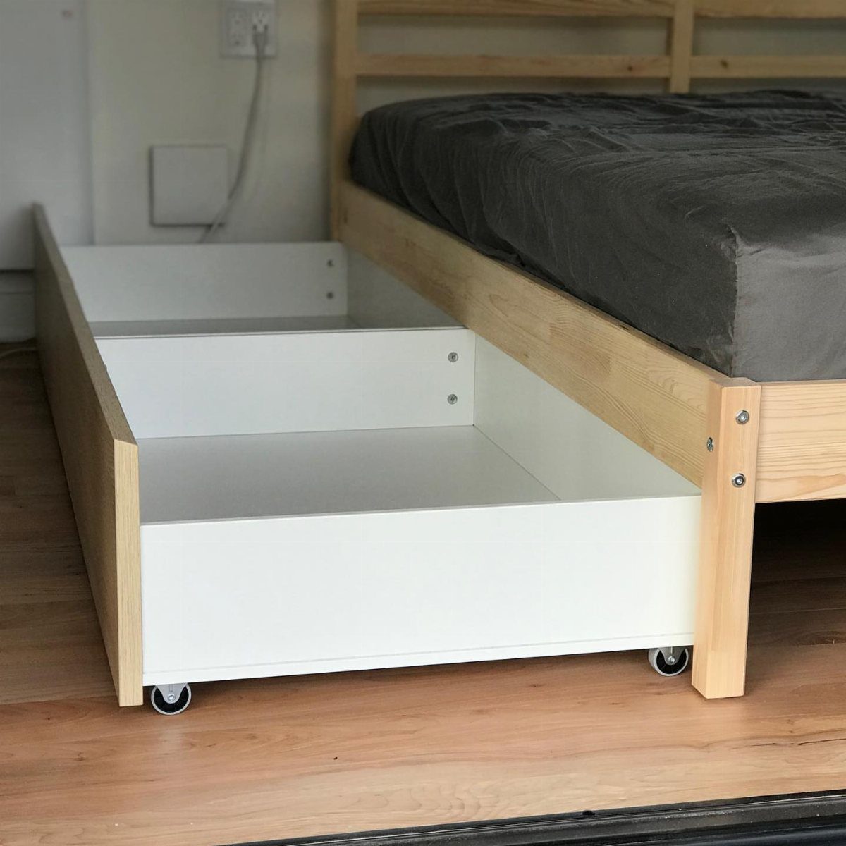 White rolling storage drawers pulled out from under a light wooden bed frame.
