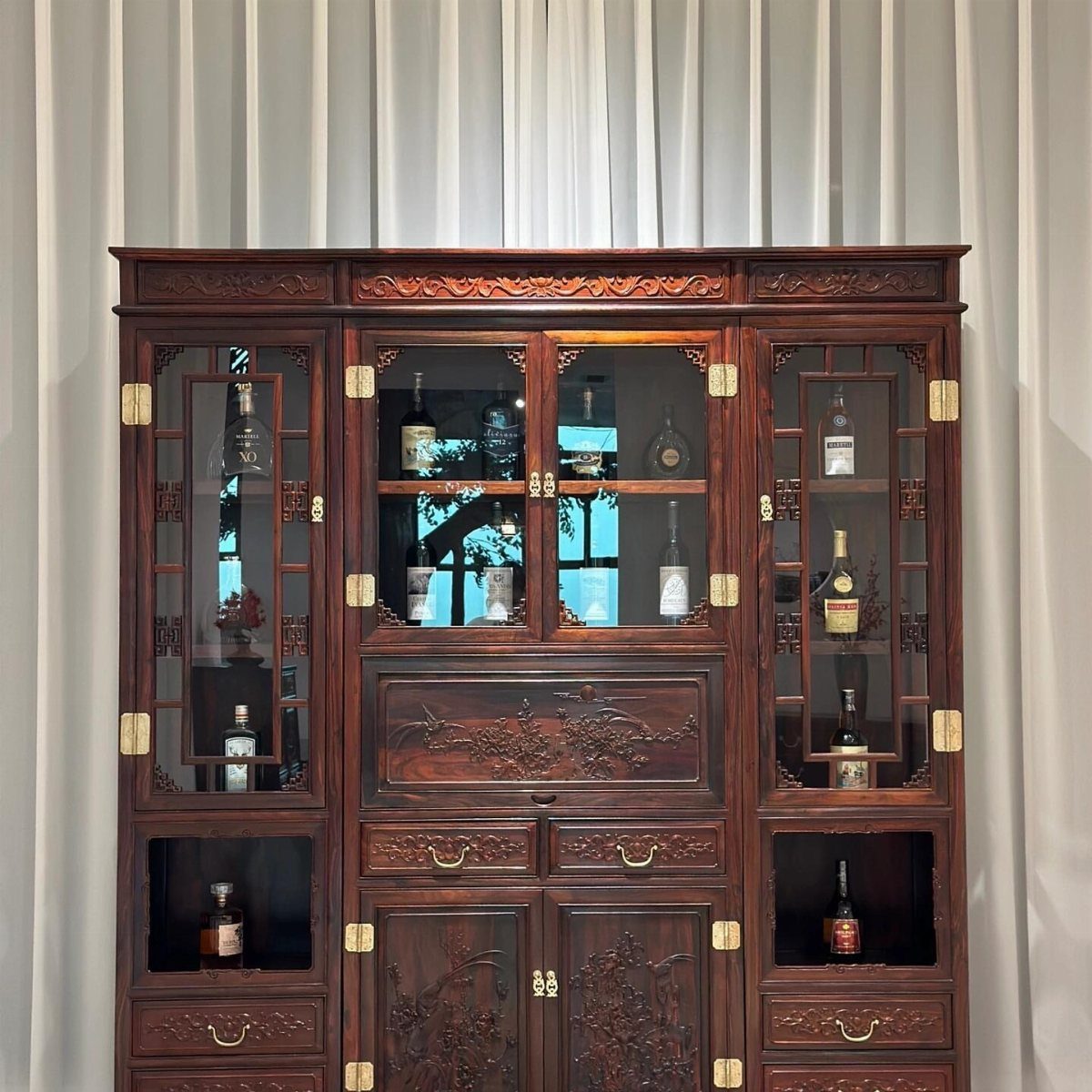 Dark wood Chinese teak cabinet with fretwork glass doors displaying various liquor bottles.