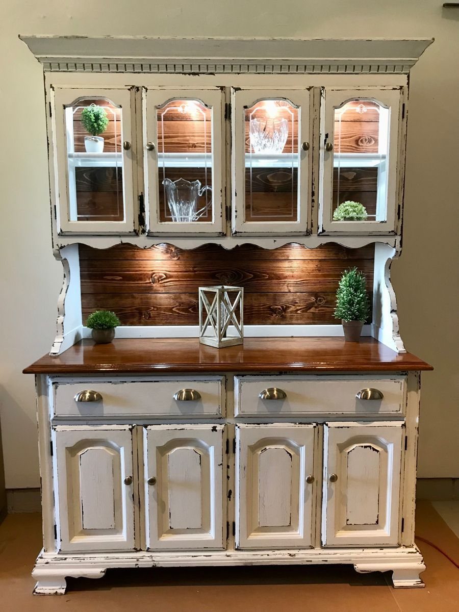 Distressed white wooden hutch with interior lighting illuminating glass cabinets and wood paneling.