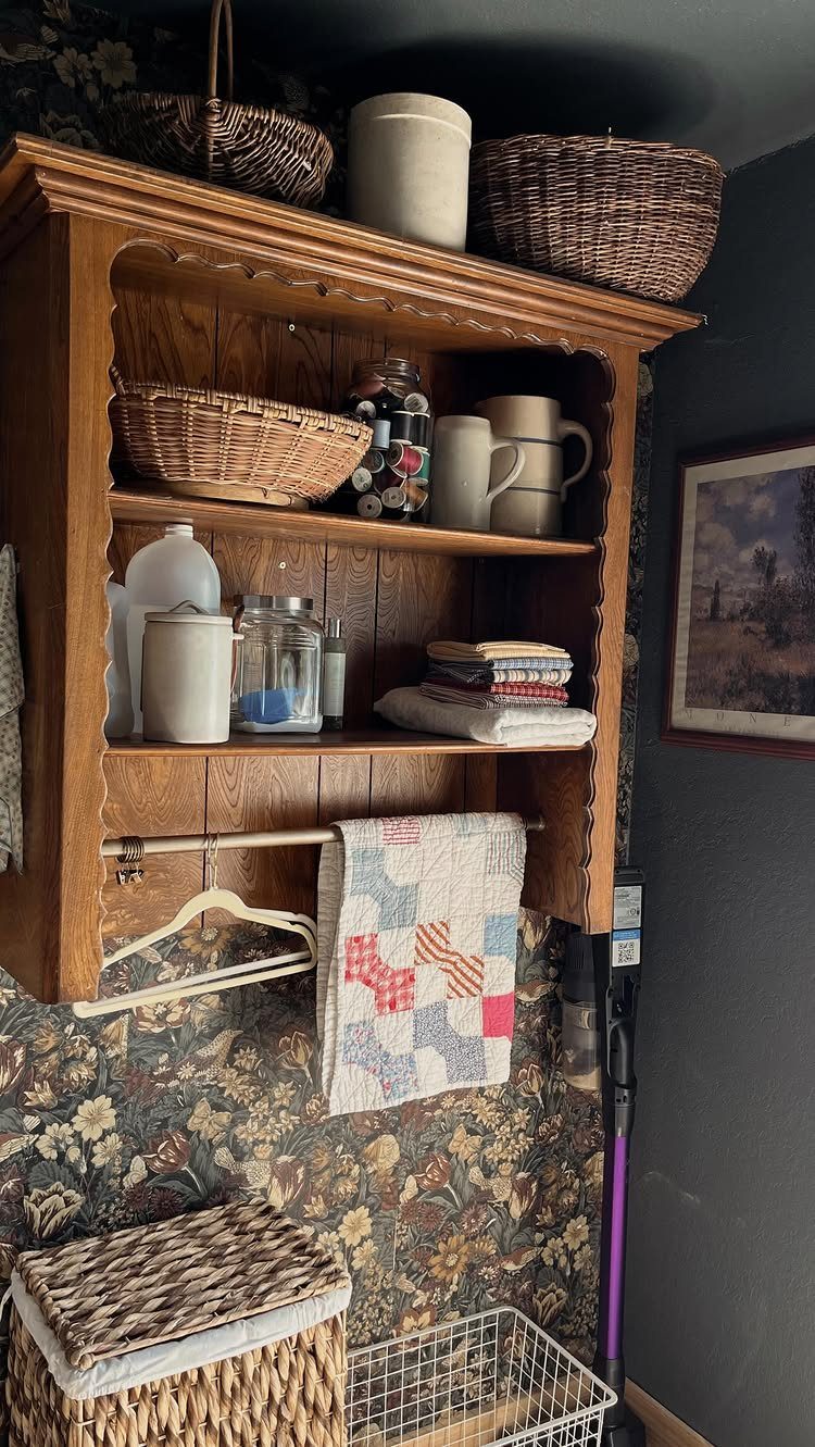 Wooden hutch top cabinet mounted on floral wallpaper holding baskets and kitchenware.
