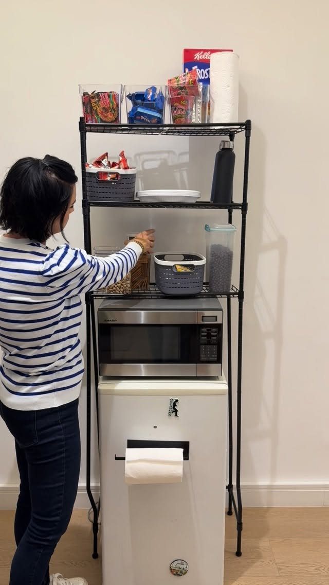 Black metal shelving unit with storage bins and microwave over a white mini-fridge.