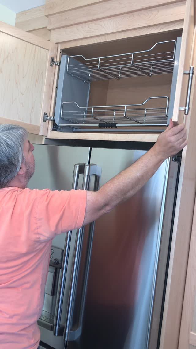 Pull-out metal wire shelves in a wooden cabinet installed above a stainless steel refrigerator.