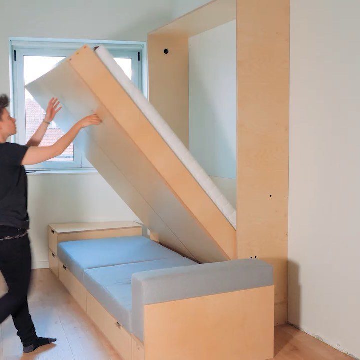 Tiny House Layout Ideas: 27 Smart Ways to Maximize Space 4 Person lifting a wooden Murphy bed into a wall cabinet above a grey sofa.